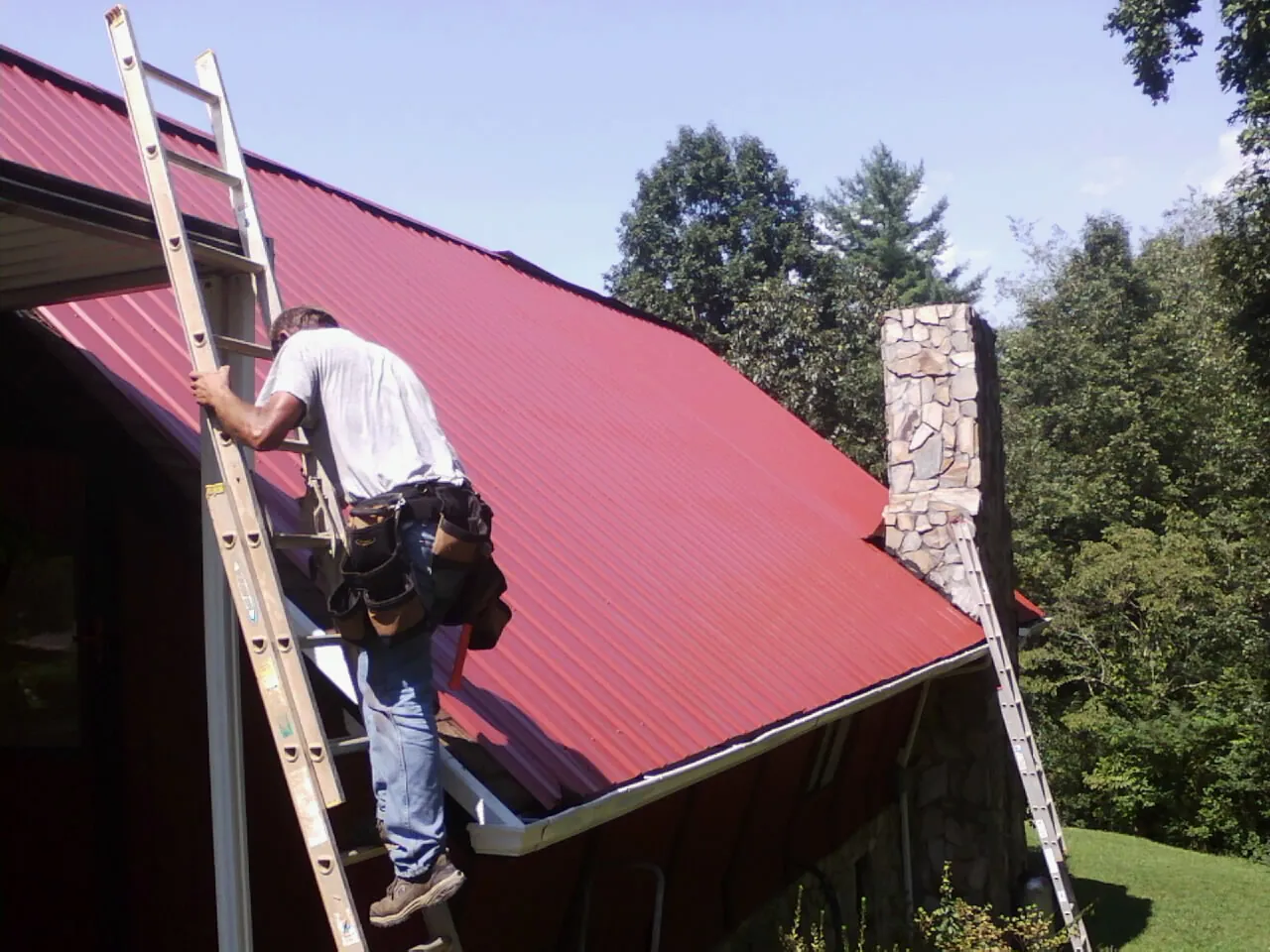 Roofer on a ladder beside a red metal roof and stone chimney — Soffit & Fascia Repair by Roofing Brothers in South Park